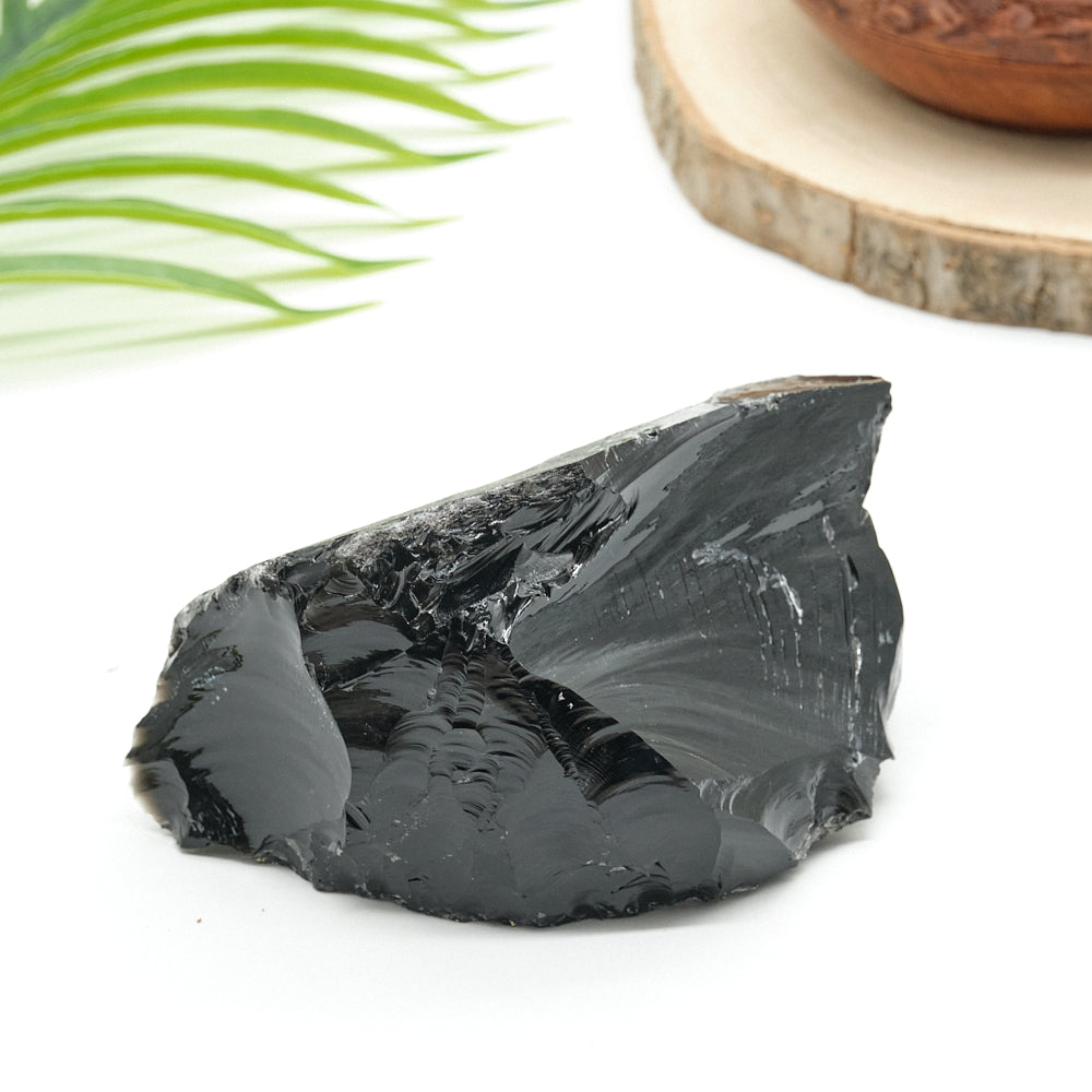 Black obsidian crystal rock stone on a white surface with a wooden bowl and green leaf in the background