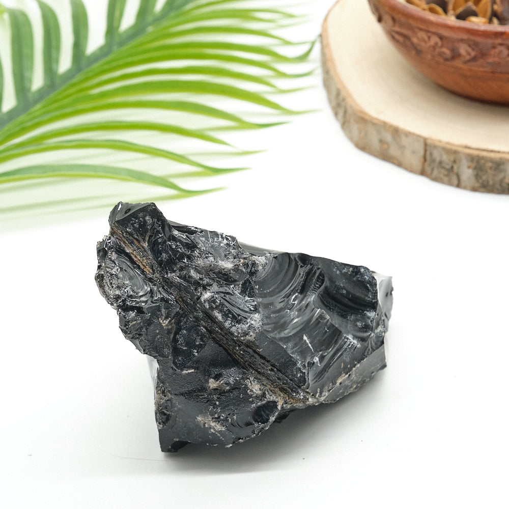 Black obsidian crystal rock on a white surface with a green leaf and wooden bowl in the background