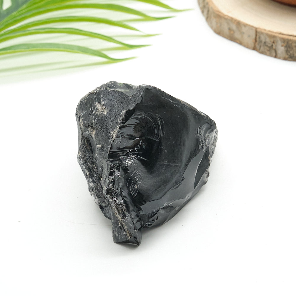 Raw black obsidian stone on a white surface with a natural wood slice and green leaves in the background