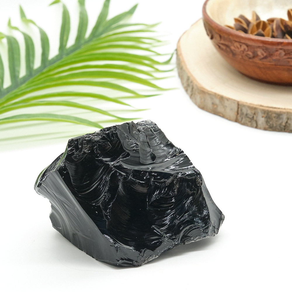 Black obsidian crystal rock on a white surface with a green leaf and wooden bowl in the background