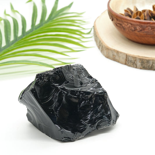 Black obsidian crystal rock on a white surface with a green leaf and wooden bowl in the background