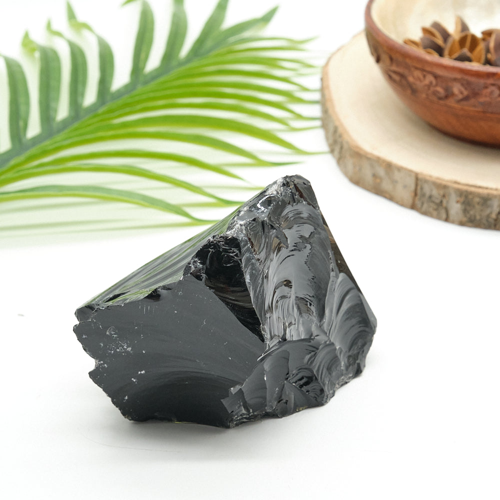 Black obsidian crystal on a white surface with green leaves and a wooden bowl in the background