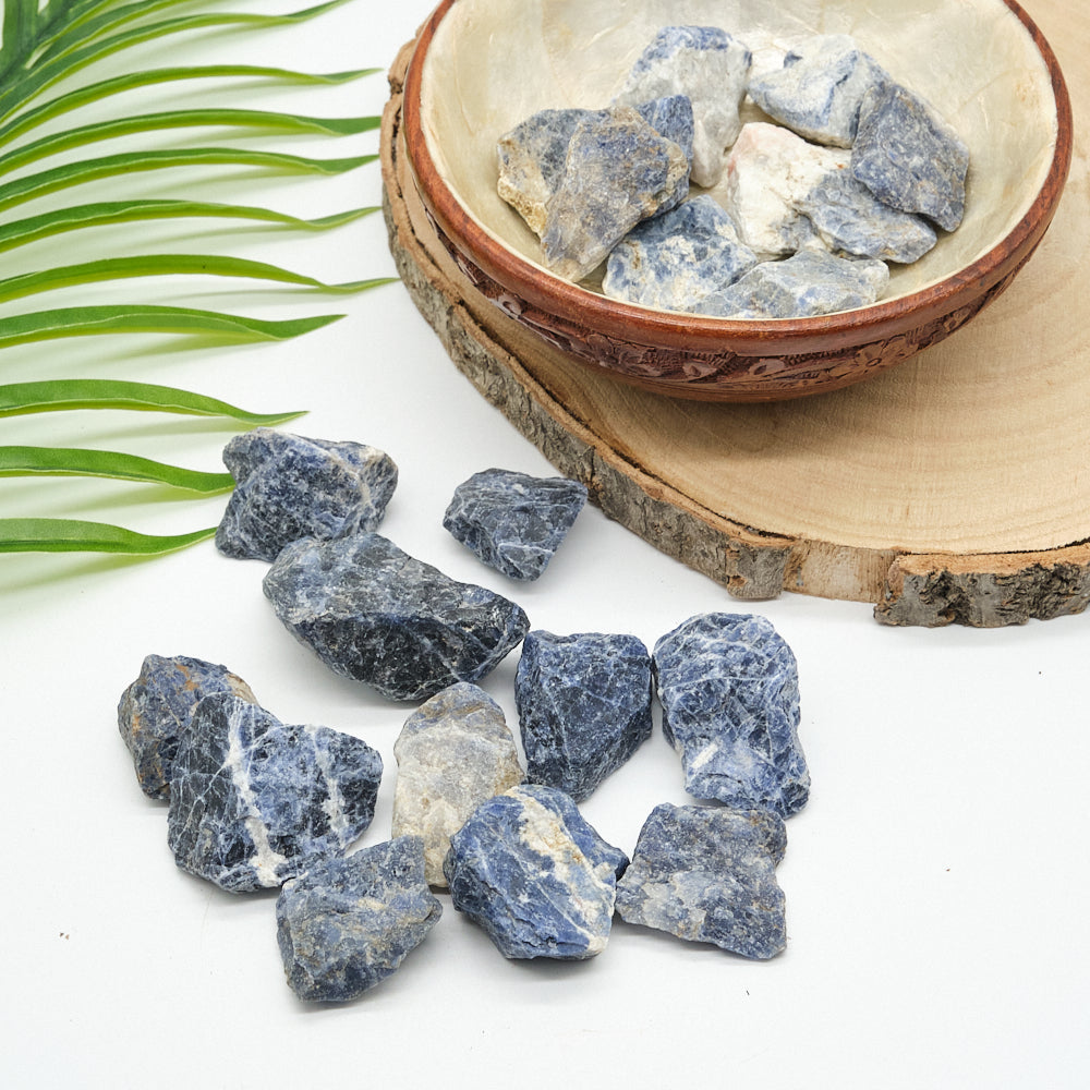 Raw sodalite stones on a white surface with a wooden bowl and green leaves.