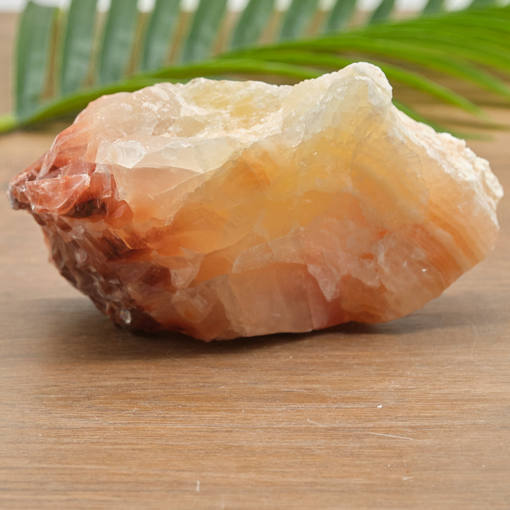 Raw yellow red calcite crystal rock on a wooden surface with green leaves in the background