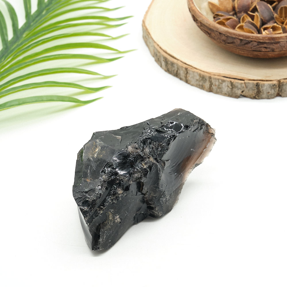 Raw black obsidian  stone on a white surface with a leaf and wooden bowl in the background