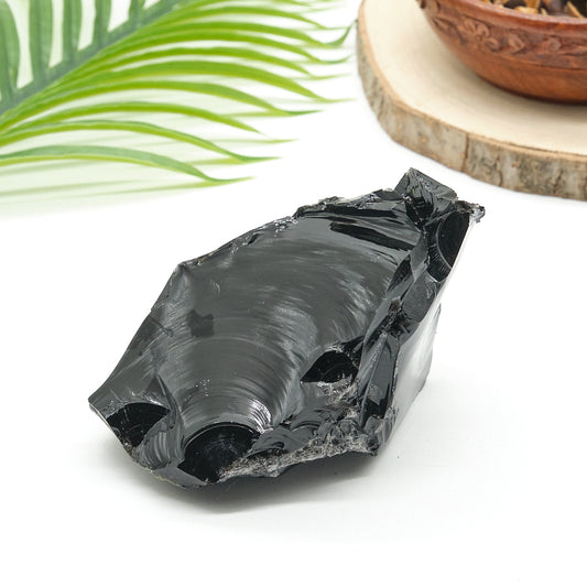 rough Black obsidian stone with a natural texture on a white surface, with green leaves and a wooden bowl in the background.