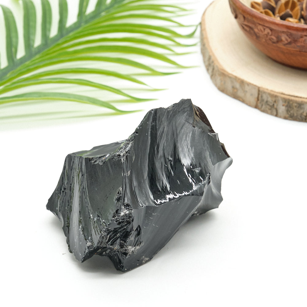 Raw obsidian crystal rock on a white surface with green leaves and a wooden bowl in the background