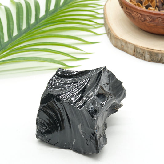 Black obsidian crystal on a white surface with a green leaf and wooden bowl in the background