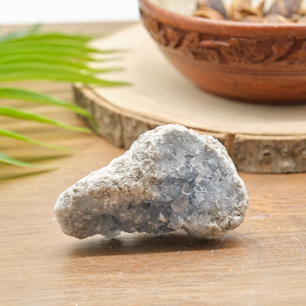 Raw celestite stone on a wooden surface with a decorative bowl and green leaves in the background
