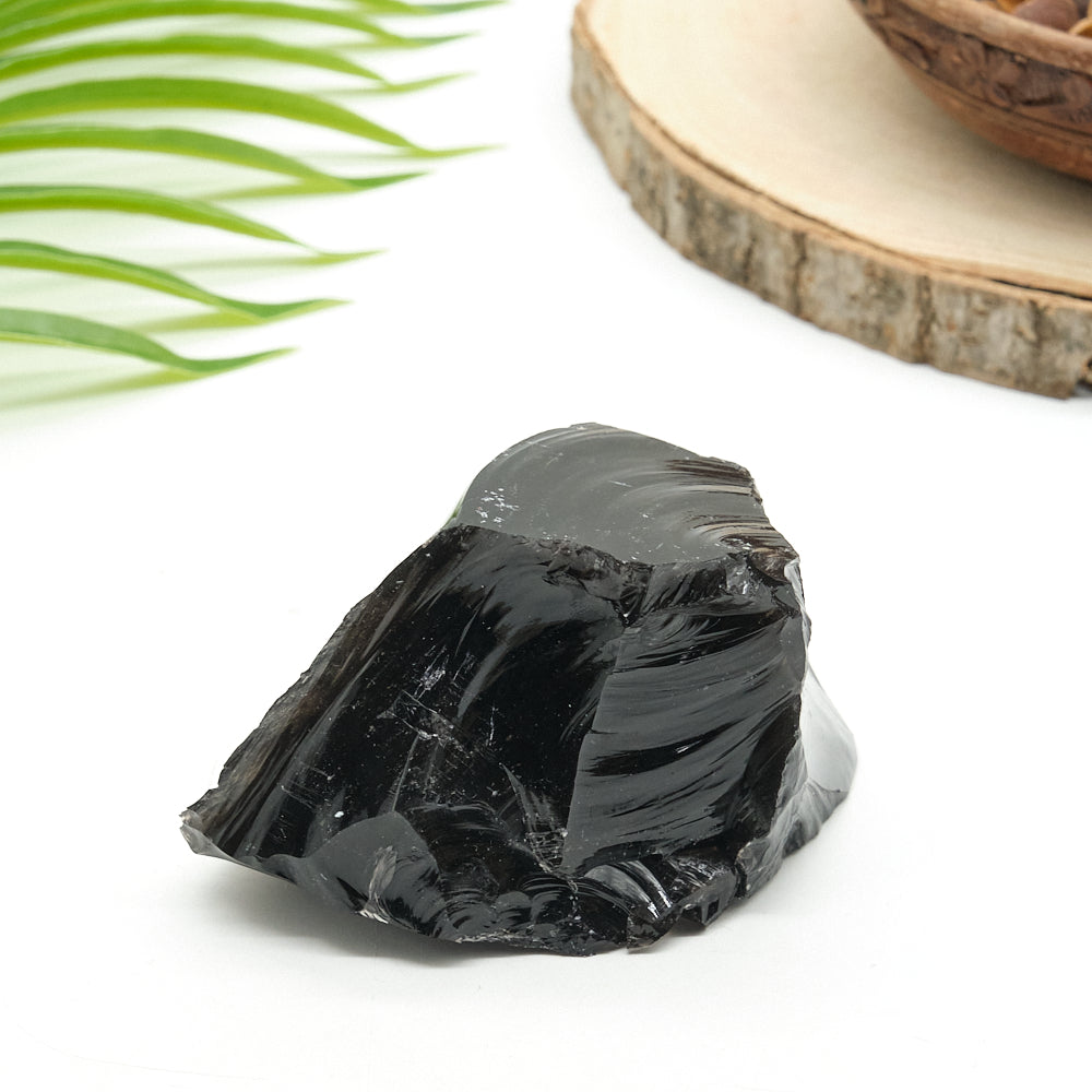Black obsidian crystal rock on a white surface with green leaves and a wooden slice in the background