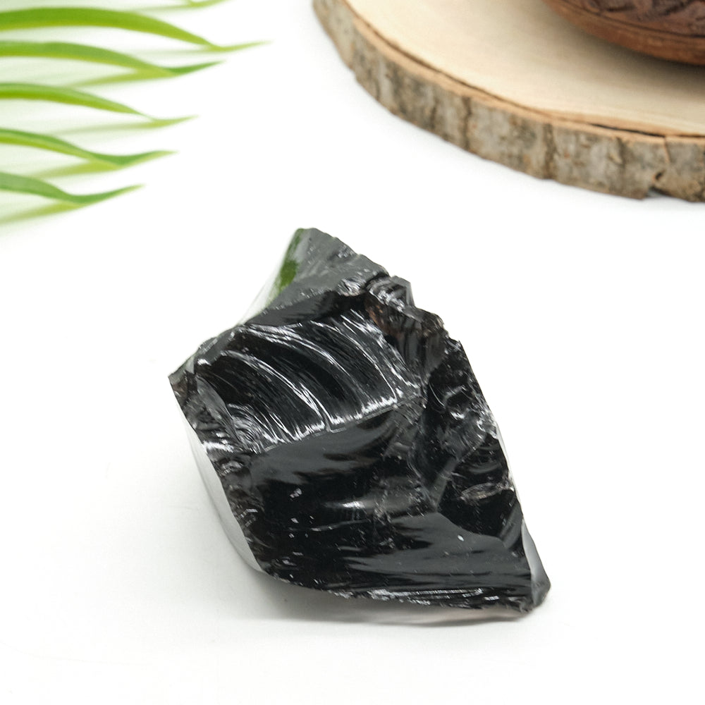 Black obsidian crystal on a white surface with green leaves and a wooden slice in the background